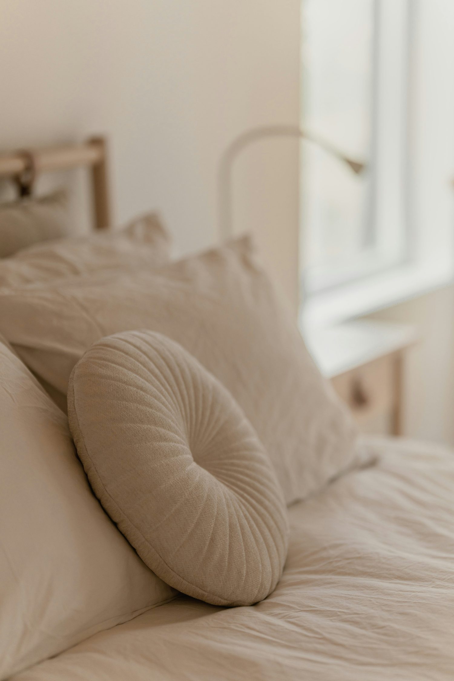 Ivory bedding with a round pleated cushion beside a bright window
