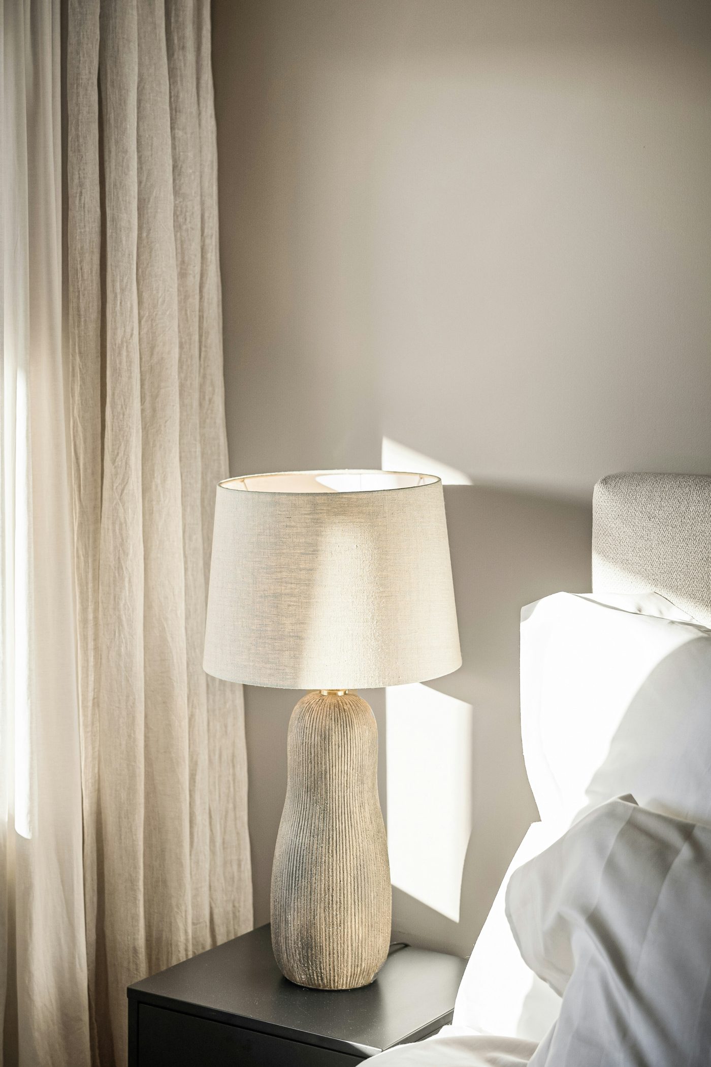 A linen-shaded table lamp beside pale curtains and white bedding