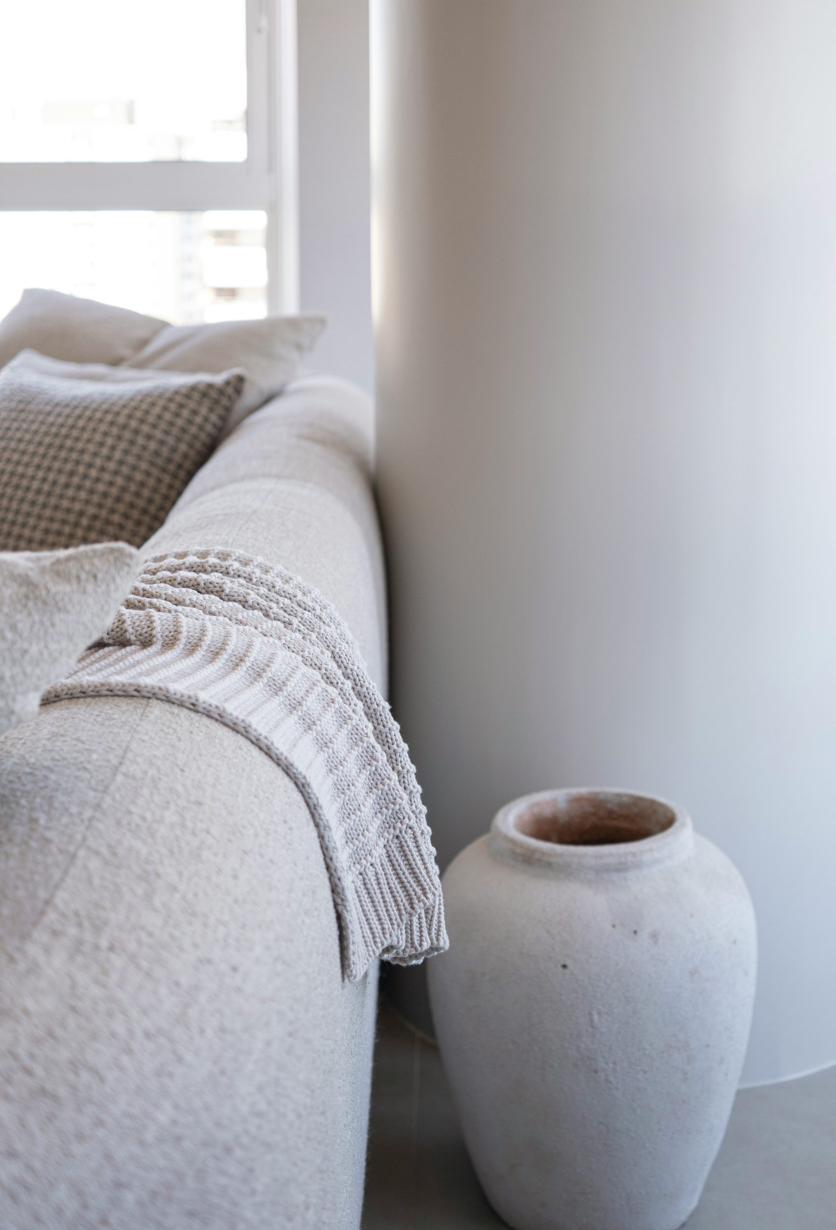 A pale linen sofa beside a tall stone-coloured ceramic vase in soft window light